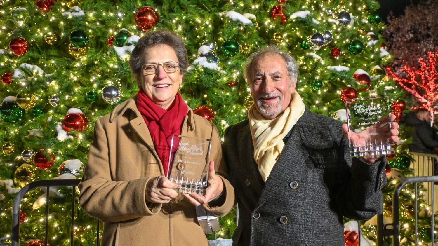 Two people holding awards in front of a large lit up Christmas tree